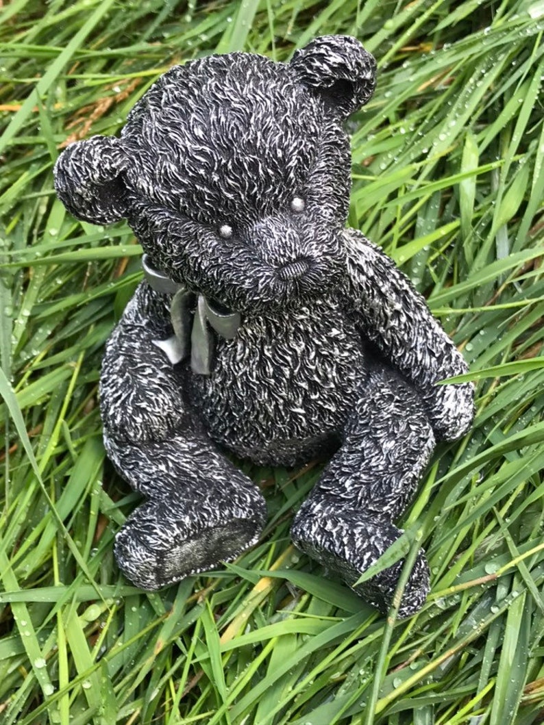 May include: A black and silver teddy bear figurine with a gray ribbon around its neck. The bear is sitting in a bed of green grass.