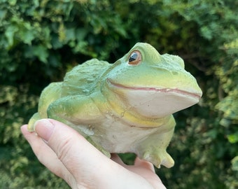 以下が含まれることがあります： 白いお腹と茶色の目をした緑色のカエルの置物。カエルは手のひらに乗っています。