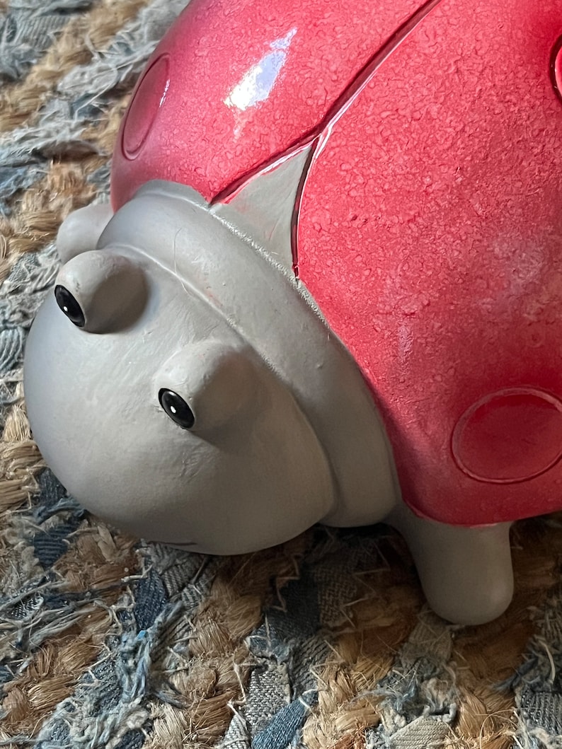 Op de afbeelding: Een close-up van een lieveheersbeestje-figuur met een rode en grijze verfbeurt. Het lieveheersbeestje heeft zwarte ogen en een getextureerd oppervlak.