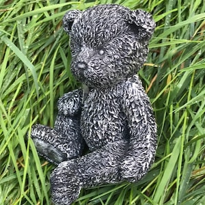 May include: A black and white teddy bear figurine sitting in a bed of green grass. The bear has a fuzzy texture and is made of a durable material.