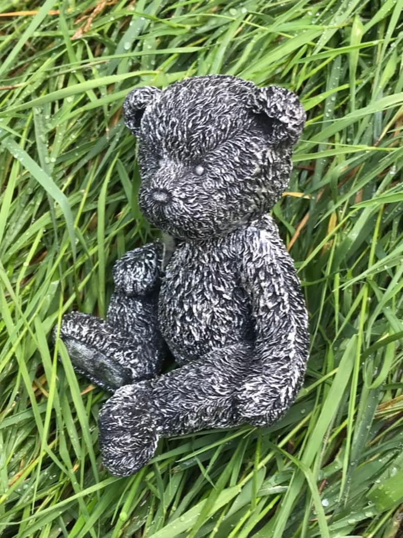 May include: A black and white teddy bear figurine sitting in a bed of green grass. The bear has a fuzzy texture and is made of a durable material.