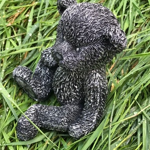 May include: A black and white ceramic teddy bear figurine sitting in a bed of green grass. The bear has a textured fur design and is sitting with its legs crossed.