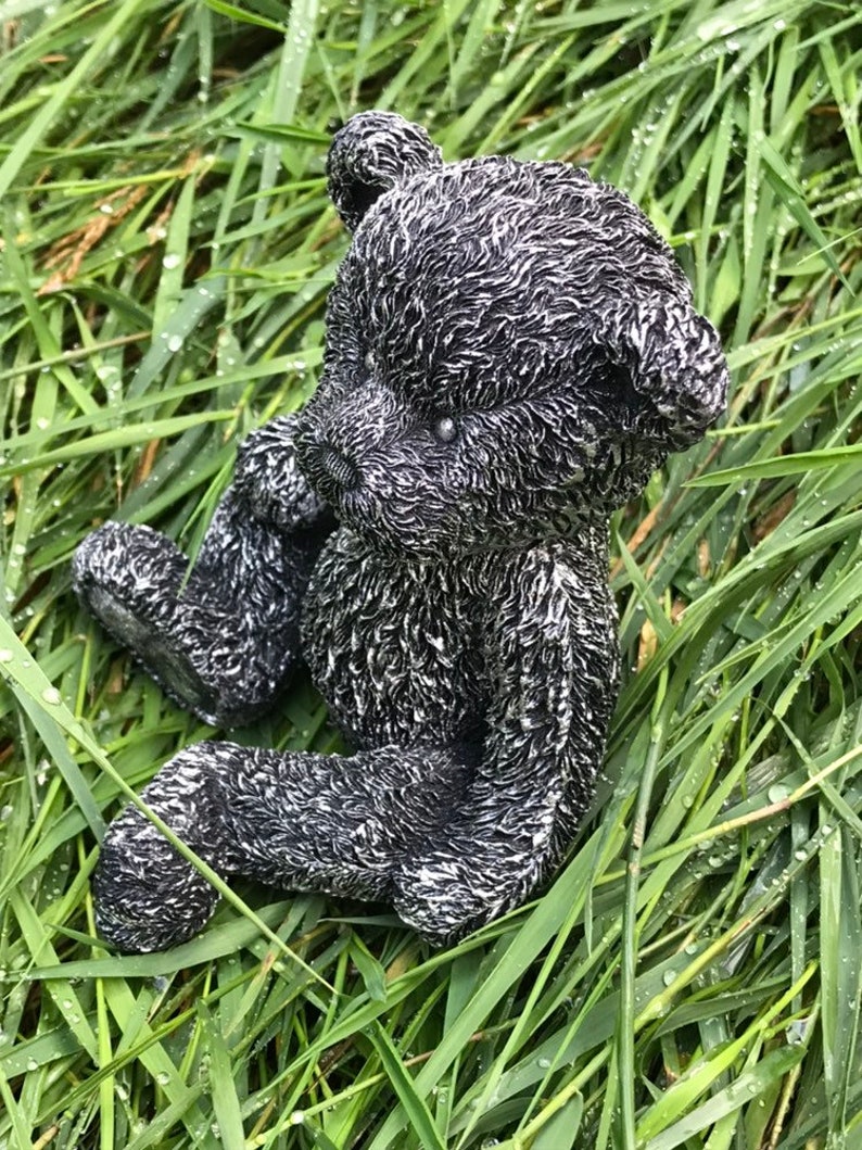 May include: A black and white ceramic teddy bear figurine sitting in a bed of green grass. The bear has a textured fur design and is sitting with its legs crossed.