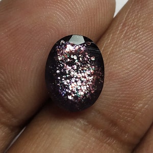 May include: An oval dark purple gemstone with shimmering, iridescent inclusions. The stone has a polished surface and a faceted top, reflecting light with a colourful shimmer. The gem is held in a hand against a white background.