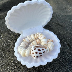 Peut inclure: Une boîte en forme de coquille blanche avec un bracelet en coquillages blanc et brun à l'intérieur. La boîte est ouverte et le bracelet est visible.