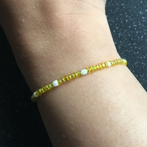 Può includere: Un braccialetto di perline con perline gialle e bianche. Il braccialetto è composto da piccole perline gialle rotonde con perline bianche quadrate. Il braccialetto è indossato su un polso.