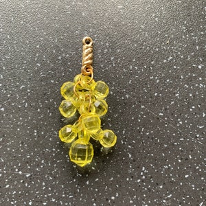Puede incluir: Un charm de uva de vidrio amarillo con un lazo de metal dorado. El charm está hecho de múltiples pequeñas cuentas de vidrio facetadas.