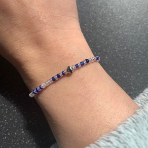 May include: A white and blue beaded bracelet with a silver charm. The bracelet is on a person's wrist.