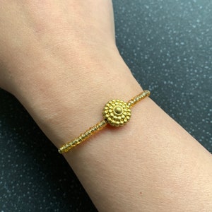 Può includere: Braccialetto dorato con piccole perline rotonde e un design centrale circolare. Il braccialetto presenta un elemento centrale decorativo con un motivo strutturato a forma di fiore. Il braccialetto è indossato su un polso.