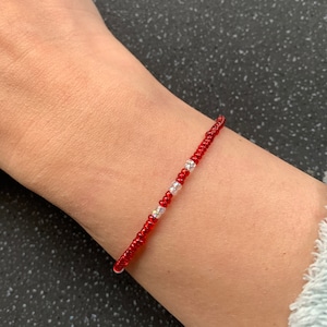 May include: A red beaded bracelet with a delicate design. The bracelet features small, vibrant red beads and a few clear, iridescent beads. The bracelet is worn on a wrist, showcasing its simple elegance and craftsmanship.