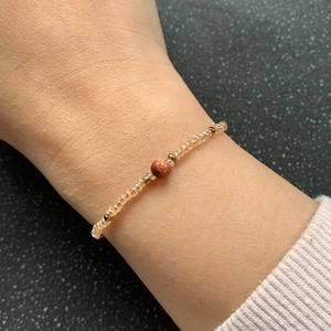 May include: A delicate bracelet made with small clear beads and a single larger red bead. The bracelet is worn on a person's wrist.