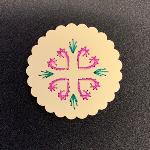 Puede incluir: Un adorno redondo de madera de color crema con un diseño bordado en rosa y turquesa. El diseño presenta cuatro corazones con una cruz en el centro.