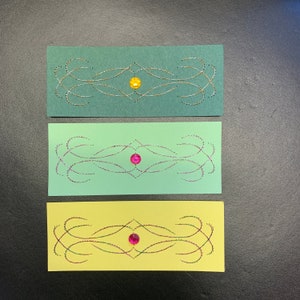 May include: Three rectangular cards with different colored backgrounds. Each card has a decorative design stitched in silver and gold thread with a single jewel embellishment in the center. The top card is green with a yellow jewel, the middle card is light green with a pink jewel, and the bottom card is yellow with a pink jewel.