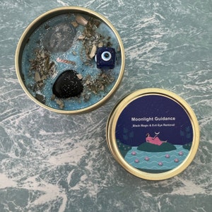 May include: A blue candle in a tin container with a black stone, clear crystal, and a blue evil eye charm. The tin has a label that reads "Moonlight Guidance Black Magic & Evil Eye Removal" with a woman in a pink dress lying on a green island in a blue lake.