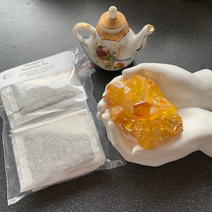 May include: Two tea bags labelled "Strength & Happiness Tea" for brewing strength, balance and wellness. The tea bags are in a clear plastic bag. A porcelain teapot with a bird design and a yellow crystal cluster are also in the image.