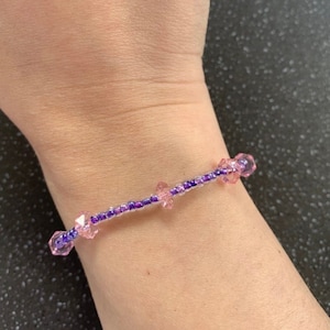 Puede incluir: Un brazalete delicado hecho con pequeñas cuentas moradas y rosadas. El brazalete está enhebrado en un hilo transparente y tiene un diseño simple.