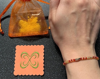 May include: A red and gold beaded bracelet with a gold charm. The bracelet is on a person's wrist. The bracelet is next to a small orange bag with yellow glitter inside and a pink card with a green design.
