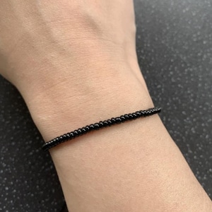 May include: A black beaded bracelet is worn on a wrist. The bracelet is made of small, round, black beads strung together. The bracelet is simple and delicate, and it sits close to the skin.