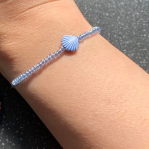 Może przedstawiać: Bransoletka z jasnoniebieskich koralików z zawieszką w kształcie muszli w kolorze białym i niebieskim.