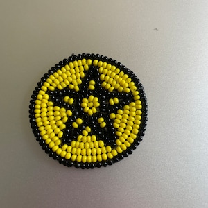 May include: A round beaded ornament featuring a black and yellow starburst design. The ornament is crafted from small, round beads that are stitched together to form a circular shape.