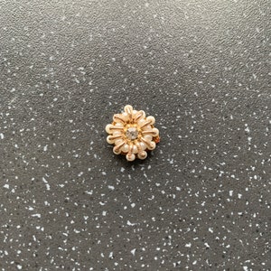 Op de afbeelding: Een kleine, goudkleurige bloemenbroche met witte parel accenten en een heldere strass steentje in het midden.