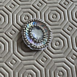 May include: A circular embroidered patch featuring a central clear oval faceted gemstone surrounded by a ring of iridescent white sequins and an outer border of small metallic silver beads, all set against a white fabric backing material.