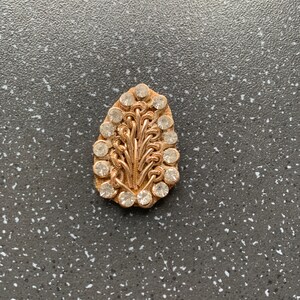 Op de afbeelding: Een gouden, traanvormige broche met een delicaat, ingewikkeld ontwerp van verweven draden en heldere strass steentjes.