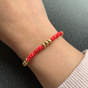 Puede incluir: Un brazalete de cuentas rojas y doradas. El brazalete está hecho con pequeñas cuentas redondas de color rojo brillante y detalles dorados.