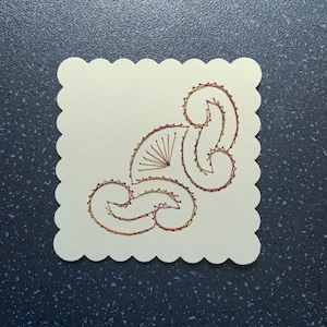 May include: A white card with a scalloped edge features a decorative design stitched in gold and red thread.