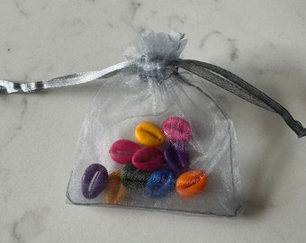 Puede incluir: Una pequeña bolsa transparente plateada con cordón llena de cuentas ovaladas de colores. Las cuentas son en tonos amarillo, rosa, rojo, morado, azul y naranja. La bolsa está atada con una cinta plateada.