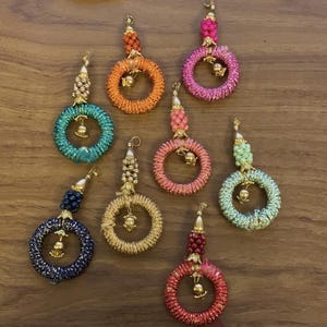 Könnte beinhalten: Sieben kreisförmige dekorative Quasten in verschiedenen Farben wie Orange, Pink, Türkis, Hellgrün, Dunkelblau, Gold und Rot. Jede Quaste besteht aus einem umwickelten Ring, einem perlenbesetzten Oberteil und einem kleinen goldenen Metallanhänger in der Mitte.