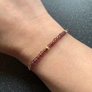 May include: A delicate bracelet made with small, dark purple beads and gold accents. The bracelet is worn on a person's wrist.