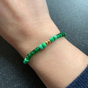 Puede incluir: Un brazalete de cuentas verdes y doradas con pequeñas cuentas redondas. El brazalete está en la muñeca de una persona.