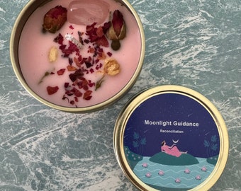 Puede incluir: Una vela rosa en una lata dorada, adornada con capullos de rosa secos, pétalos y un cristal transparente. La tapa presenta un diseño azul con las palabras "Moonlight Guidance" y "Reconciliation".