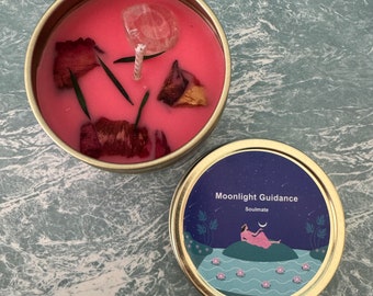 Puede incluir: Una vela rosa en un recipiente de hojalata con un cristal transparente y pétalos de rosa secos. La tapa de la lata tiene una ilustración azul y blanca de una mujer sentada en una roca en un lago con el texto "Moonlight Guidance Soulmate".
