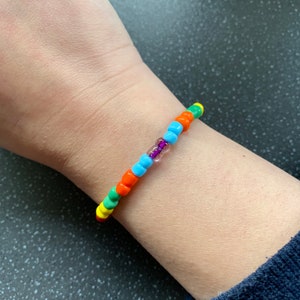 Op de afbeelding: Een kleurrijke kralen armband met afwisselende oranje, blauwe, groene en gele kralen. Een enkele paarse kraal bevindt zich in het midden van de armband.