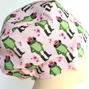 May include: A light pink surgical scrub cap featuring a repeating pattern of green frogs wearing cowboy hats and boots. The frogs are interspersed with pink starburst designs. The cap is made of a soft, likely cotton, fabric.