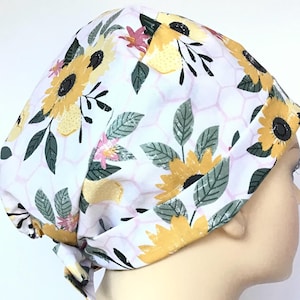 May include: A white scrub hat with a sunflower and honeycomb pattern. The hat has a tie at the back and a button closure at the top.
