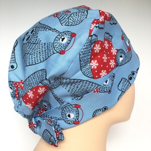 May include: A light blue scrub cap featuring a repeating pattern of illustrated bears. The bears are depicted in black line art with red accents, wearing red sweaters adorned with white snowflake patterns. The cap has a tie-back closure.