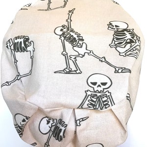 Op de afbeelding: Een beige scrubmuts van stof met een herhalend patroon van zwarte skeletten in verschillende yogaposes.
