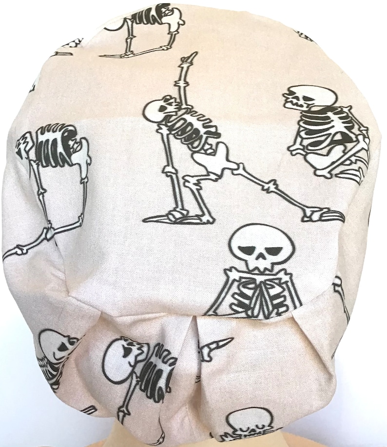 Op de afbeelding: Een beige scrubmuts van stof met een herhalend patroon van zwarte skeletten in verschillende yogaposes.