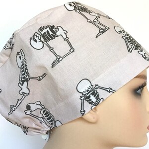 Op de afbeelding: Een beige scrub-muts met een zwart skeletpatroon. De muts heeft een strik aan de achterkant.