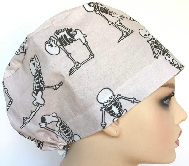 Op de afbeelding: Een beige scrub-muts met een zwart skeletpatroon. De muts heeft een strik aan de achterkant.