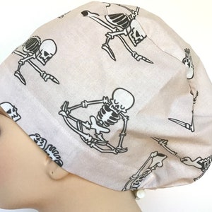 Op de afbeelding: Een lichtbeige scrubmuts met een herhalend patroon van zwarte skeletten in verschillende yogaposes.