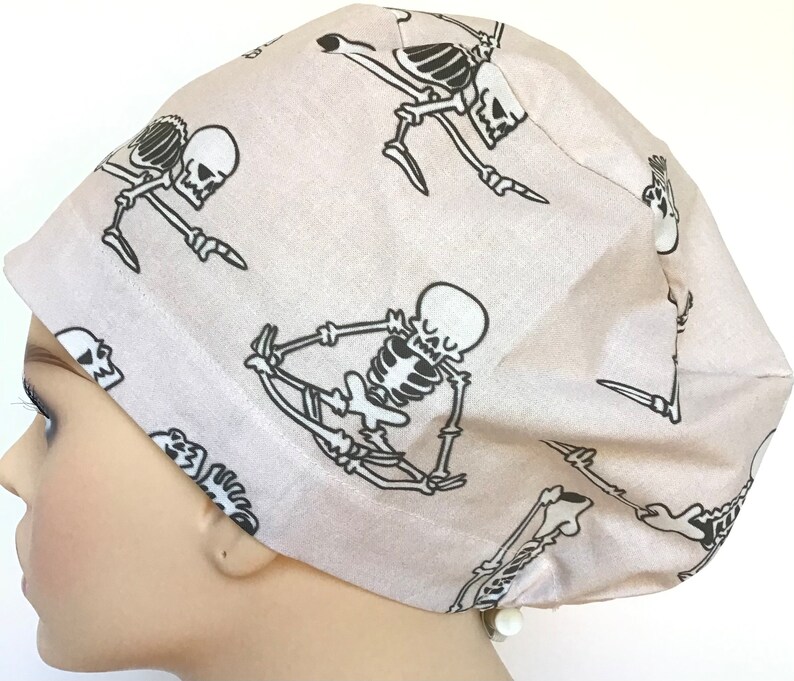 Op de afbeelding: Een lichtbeige scrubmuts met een herhalend patroon van zwarte skeletten in verschillende yogaposes.