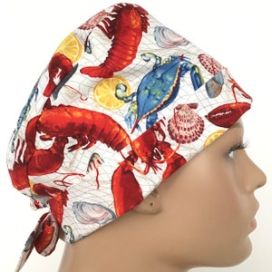 May include: A white scrub hat with a colorful seafood print featuring lobsters, crabs, and seashells. The hat has a tie at the back.