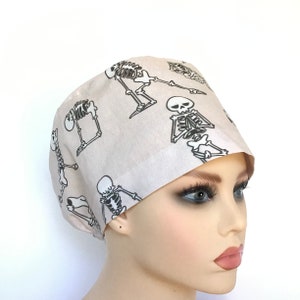 Op de afbeelding: Een beige scrub-muts met een herhalend patroon van zwarte skeletten. De skeletten staan in verschillende poses, sommige staan, sommige zitten en sommige houden hun schedel vast.