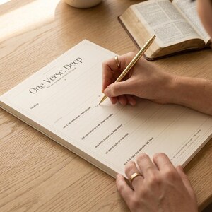 May include: A cream-coloured paper notepad titled One Verse. Deep. with printed prompts for scripture study, including lines for writing verses, reflections, and prayers. A gold-toned metal pen rests on the page, and an open book is visible nearby.