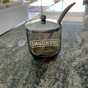 May include: A glass jar with a silver lid and a wooden spoon. The jar is filled with a dark liquid and has the word "Smucker's" printed on the side.