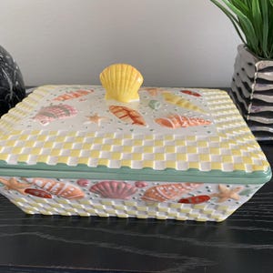 May include: A ceramic rectangular serving dish with a lid. The dish is decorated with a yellow and white checkered pattern and seashells in various colors. The lid has a seashell knob.